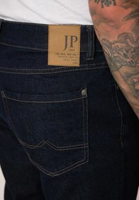 Tummat farkut, joissa on ruskea nahkapaneeli, jossa on "JP 1880" -logo, näkyvää ommelta ja takatasku, jossa on koristeellinen sauma.