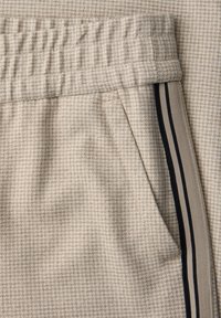 Pantalones de tela beige con pequeño patrón cuadriculado, cintura elástica, bolsillo lateral y una franja beige con dos líneas negras delgadas en la costura lateral.