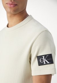 Světle béžové bavlněné tričko s krátkým rukávem a jemnou strukturou. Na rukávu se nachází černá nášivka s bílým logem "Calvin Klein Jeans".