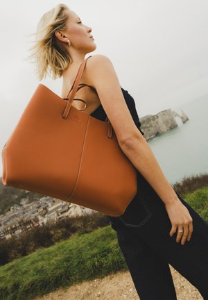 Mujer rubia con vestido negro que lleva un gran bolso tote de cuero marrón, de pie en una colina con hierba con vistas a acantilados costeros y un pueblo.