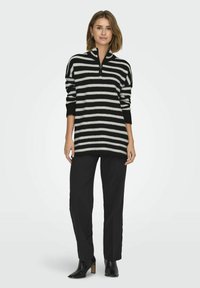 Zwart-wit gestreepte gebreide trui met een halve rits kraag. Het model draagt op maat gemaakte zwarte broek en zwarte haklaarzen.