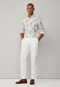 Pantalones largos blancos de corte relajado combinados con una camisa de manga corta con estampados de palmeras verdes sobre un fondo claro. Zapatos de cuero marrón.