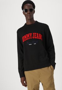 Svart strikkegenser med rød "TOMMY JEANS"-innskrift og blå aksent. Ribbekanter på mansjettene og nederkanten, sammen med khaki-bukser. Solbriller brukt.