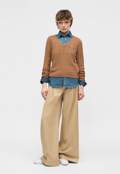 Personne portant un pull marron en maille torsadée par-dessus une chemise en denim bleu, un pantalon beige à jambes larges et des baskets blanches, debout devant un fond uni.