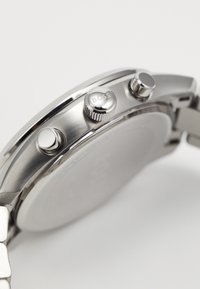 Ασημί ρολόι χειρός από ανοξείδωτο ατσάλι με υφή στη στεφάνη, κορώνα με χαραγμένο λογότυπο και κουμπιά. Διαθέτει επίπεδη, λεία πλάτη από καπάκι.