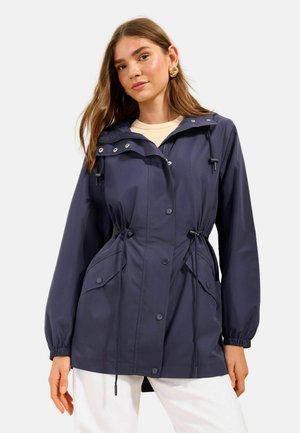 Žena v tmavomodrej nepremokavej bunde s kapucňou so šnúrkami v páse a vreckami, v kombinácii s bielymi nohavicami a zlatými kruhovými náušnicami.