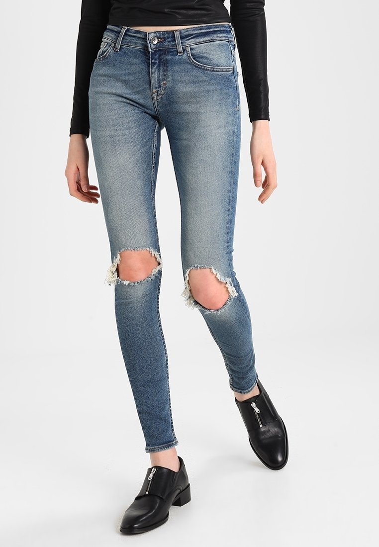 Slim-fit niebieskie dżinsy z dużymi dziurami na obu kolanach, noszone z czarnymi botkami na zamek do kostek i czarną bluzką z długim rękawem.