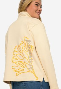Sudadera de color crema con un corte relajado, que presenta un gran diseño de hoja amarilla y texto "cosido" en gris en la parte posterior. Textura suave, diseño minimalista.