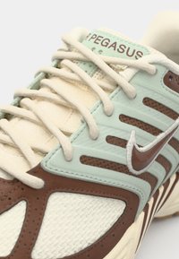 Les sneakers Nike Pegasus présentent un mélange d'accents vert menthe clair et marron, une tige en mesh, une semelle en caoutchouc texturée et des lacets beiges avec un logo sur la languette.