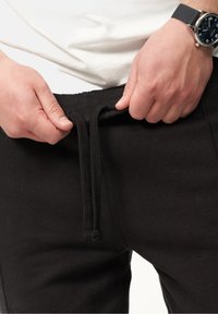 Schwarze Jogginghosen mit einem elastischen Bündchen und Kordelzug. Glatte Stoffstruktur. Hände, die das Bündchen anpassen. Uhr am Handgelenk sichtbar.