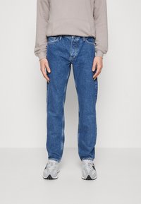 Vaqueros de mezclilla azul con un corte recto, que presentan costuras en contraste y un diseño clásico de cinco bolsillos, combinados con una sudadera gris claro y zapatillas deportivas.
