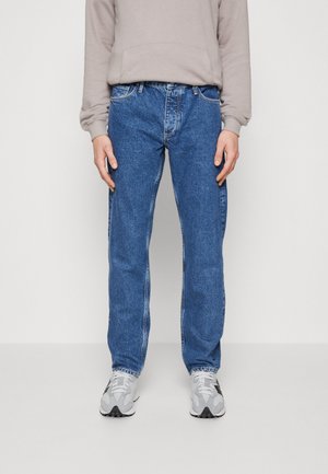 Vaqueros de mezclilla azul con un corte recto, que presentan costuras en contraste y un diseño clásico de cinco bolsillos, combinados con una sudadera gris claro y zapatillas deportivas.