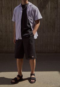 Camicetta corta viola chiaro, t-shirt nera, pantaloni corti neri con patch logo e sandali neri con cinghie testurizzate. Abbigliamento casual.