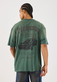 Mand med fletninger iført en grøn oversized T-shirt med et vintage bilgrafik og tekst relateret til Los Angeles og motorvejs tuning.
