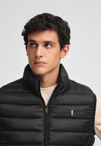 Svart vadderad väst med hög krage, dragkedjestängning och guldlogotyp som accent. Har ett quiltat mönster, slät textur, bärs över ett beige plagg.