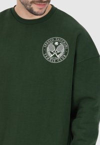 Felpa verde con un logo rotondo che presenta racchette da tennis, foglie e testo. Realizzata in tessuto morbido, con una vestibilità comoda e polsini a coste.
