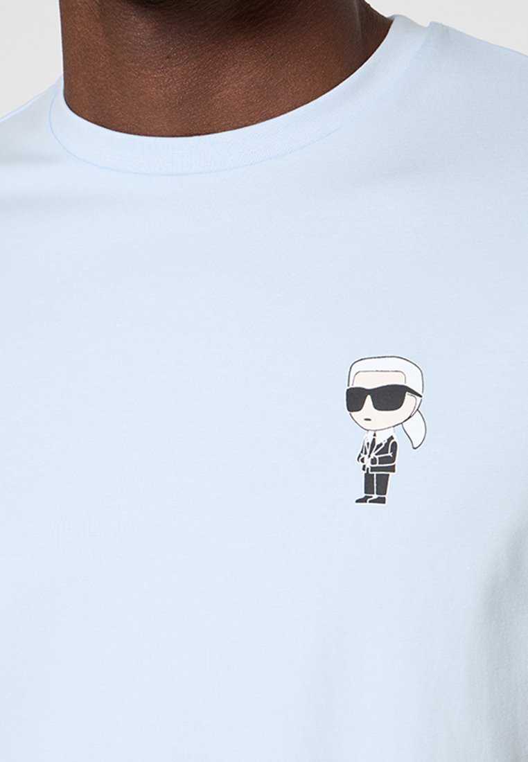 Pequeña figura de dibujo animado con cabello blanco recogido, gafas de sol negras y un traje negro en la parte superior del pecho de una camisa azul claro.