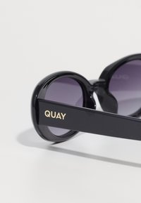 Óculos de sol oversized pretos com lentes roxas escuras. As hastes têm um acabamento brilhante com "QUAY" em letras douradas. Textura suave em toda a peça.