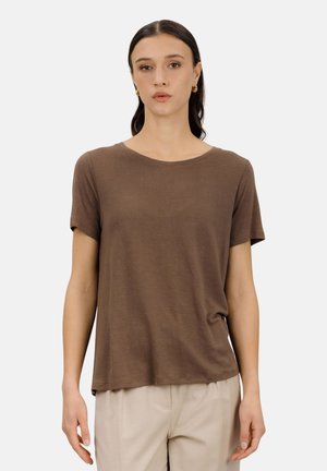 Ung kvinne med mørkt hår iført gulløreringer, en brun kortermet skjorte og beige bukser, stående foran en ensfarget bakgrunn.