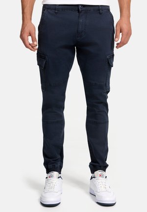 Uomo che indossa pantaloni jogger cargo blu navy con polsini elasticizzati e sneakers bianche Reebok, in piedi contro uno sfondo semplice.