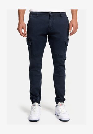 Uomo che indossa pantaloni jogger cargo blu navy con polsini elasticizzati e sneakers bianche Reebok, in piedi contro uno sfondo semplice.