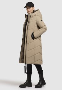 Beige quiltet lang jakke med hætte, med sorte detaljer og lynlås. Bæres over en sort top og parret med sorte støvler.