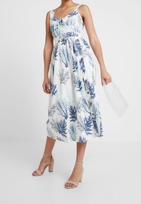 Femme portant une robe midi sans manches blanche avec un motif d'algues marines bleues et des boutons sur le côté, des sandales à talons nus, tenant un sac à main en perles transparentes.