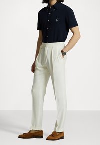 Marinblå kortärmad polo med vitt logotyp, hög midja vita veckade byxor och bruna mocka loafers. Klocka på handleden ger extra detalj.