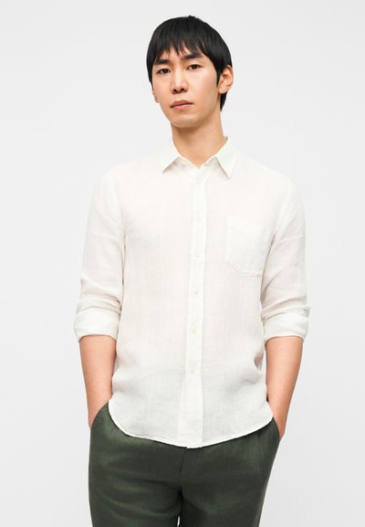 Ελαφρύ λευκό λινό πουκάμισο με γιακά με κουμπιά, μία τσέπη στο στήθος και μανίκια γυρισμένα. Συνδυασμένο με σκούρο πράσινο παντελόνι.