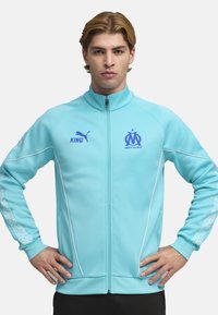 Chaqueta deportiva turquesa con cremallera al frente, cuello alto, detalles en blanco y mangas con estampado. Logotipos en azul en el pecho y el brazo derecho.