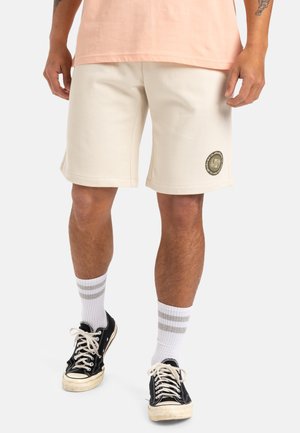 Pantalones cortos de color crema con un corte relajado, presentan un parche redondo de logo en el muslo izquierdo, combinados con zapatillas negras y calcetines blancos con rayas grises.