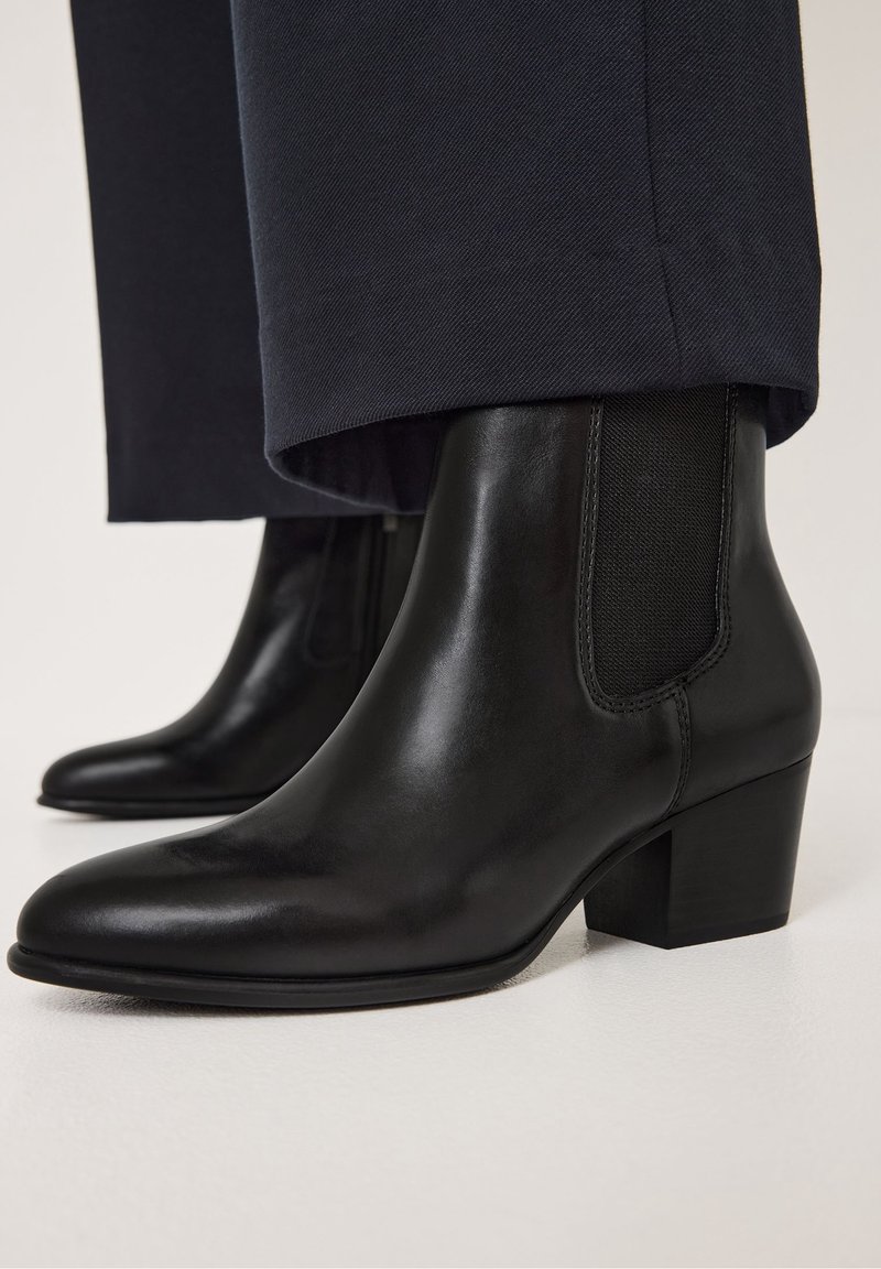 Bottines en cuir noir à talons blocs portées avec un pantalon foncé, montrées en gros plan sous un angle latéral bas.