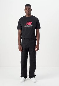Fekete rövid ujjú póló rózsaszín New Balance logóval, fekete nadrággal és fehér sportcipővel párosítva, a modell egyenes testtartásban áll, egyszínű háttér előtt.