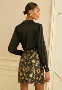 Musta pitkähihainen pusero yhdistettynä vartalonmyötäiseen, kullankuvioituun hameeseen, jossa on taivaankappaleiden kuvia. Sileä pinta, jossa näkyvät saumat ja napit.