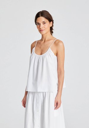 Weiße Baumwoll-Camisole mit dünnen, verstellbaren Trägern, gerafftem Halsauschnitt und lockerer Passform. Kombiniert mit einem weißen knöpfbaren Rock.
