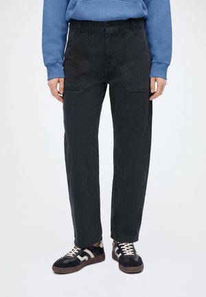 Person iført sorte bukser med lige ben med frontlommer, blå sweatshirt og sorte sneakers med hvide detaljer og brune såler.