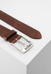 Cinturón de cuero marrón con una hebilla de metal plateado, que presenta el nombre de la marca "Levi's", y seis agujeros de ajuste para el tamaño.