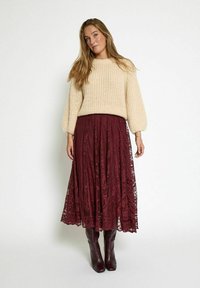 Beżowy sweter dzianinowy z balonowymi rękawami, zestawiony z burgundową koronkową spódnicą maxi. Modelka ma na sobie burgundowe buty za kolano.