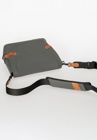 Graue Leder-Crossbody-Tasche mit braunen Lederakzenten, rechteckiger Form, verstellbarem Schulterriemen und einem geprägten Logo auf der Vorderseite.