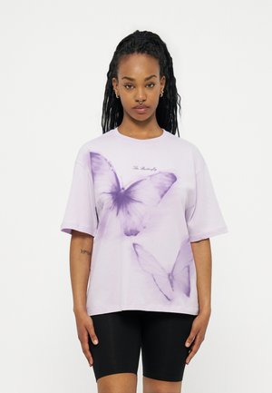 Νεαρή γυναίκα με μακριά πλεξούδα, ντυμένη με ένα ανοιχτό μωβ φαρδύ T-shirt με μεγάλα μοτίβα μοβ πεταλούδας και μαύρο σορτς για ποδηλασία.