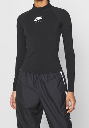 Person trägt ein schwarzes langärmeliges Nike Air Top und schwarze Nike Trainingshose mit weißen Streifen, steht vor einem grauen Hintergrund.