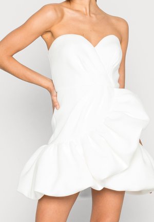 Biała sukienka bez ramiączek z dekoltem w kształcie serca i obszerną, falbaną spódnicą, prezentowana przez osobę z jedną ręką na biodrze.