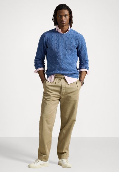 Μπλε πουλόβερ με πλέξη κασμίρ, φορεμένο πάνω από ελαφρύ ροζ πουκάμισο, συνδυασμένο με καμηλό παντελόνι και λευκά αθλητικά παπούτσια. Χαλαρός, άνετος στυλ.