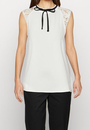 Ærmeløs hvid bluse med blonder korte ærmer og sort sløjfe ved kraven, kombineret med sorte bukser på en stående person.