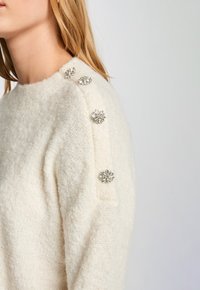 Valkoinen neulepusero, jossa on tekstuuripintainen viimeistely ja koristeelliset kristallikukkasovitteet olkapäillä. Pyöreä kaula-aukko.