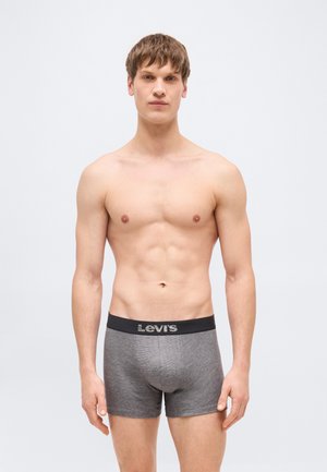 Jauns vīrietis stāv ar seju pret priekšu, valkā pelēkas Levi's bokserbikses ar melnu jostas daļu, vienkāršs gaišs fons.