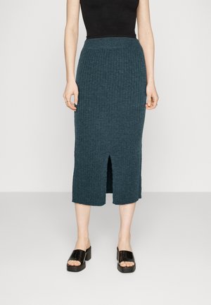 Strick-Midi-Rock in Teal mit gerippter Textur, seitlichem Schlitz und eng anliegendem Bund, kombiniert mit schwarzen Sandalen mit Blockabsatz und offenen Zehen.