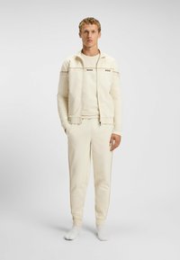 Lichtcrème trainingsoutfit gemaakt van zacht materiaal. Bevat een zip-up jack, bijpassende broek met zijkanten en het woord "BOSS" op de borst.