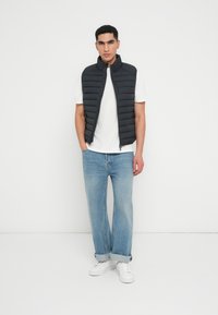 Sort polstret vest med høj krave over en hvid t-shirt, parret med lyseblå lige bukser og hvide sneakers. Hvid baggrund.