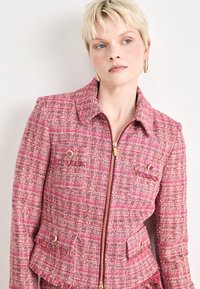 Vaaleanpunainen tweed-takki, jossa on vetoketju edessä, terävä kaulus ja kaksi rintataskua. Varustettu kultasävytteisillä yksityiskohdilla ja teksturoidulla pinnalla, jossa on frangeja.